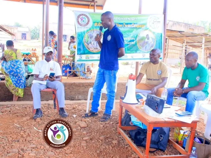 A speaker addresses a community gathering beside a Sustain Rise banner