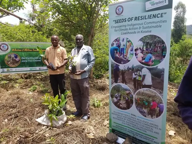 Two men hold seedlings next to the Seeds of Resilience banner